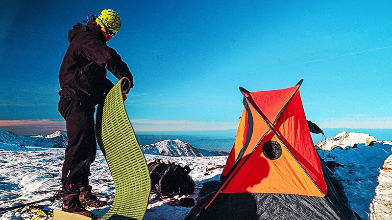 On a mountain spot in winter, a camper arranges his sleeping pad near his orange tent